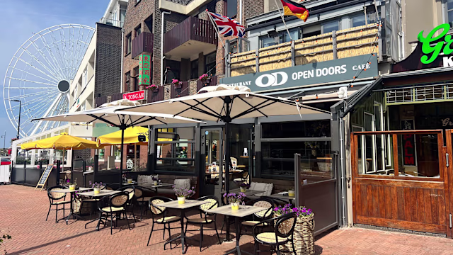 Het beeld toont een levendige restaurantgevel met buitenzitplaatsen, versierd met kleine bloemstukken. De parasols bieden schaduw voor de gasten en de algehele sfeer is uitnodigend en informeel. Op de achtergrond voegt een groot reuzenrad een speels element toe aan de omgeving, wat de levendige ambiance versterkt. De signage van het restaurant is duidelijk en geeft aan dat het een gastvrije plek is.