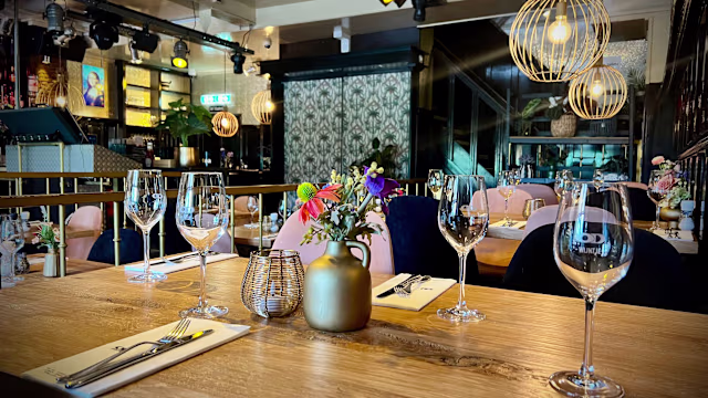 Stijlvol restaurantinterieur met een mix van moderne en rustieke elementen, houten tafels met elegante glazen en netjes gerangschikt bestek, zachte verlichting van decoratieve hanglampen en bloemstukken die kleur toevoegen aan de uitnodigende sfeer.