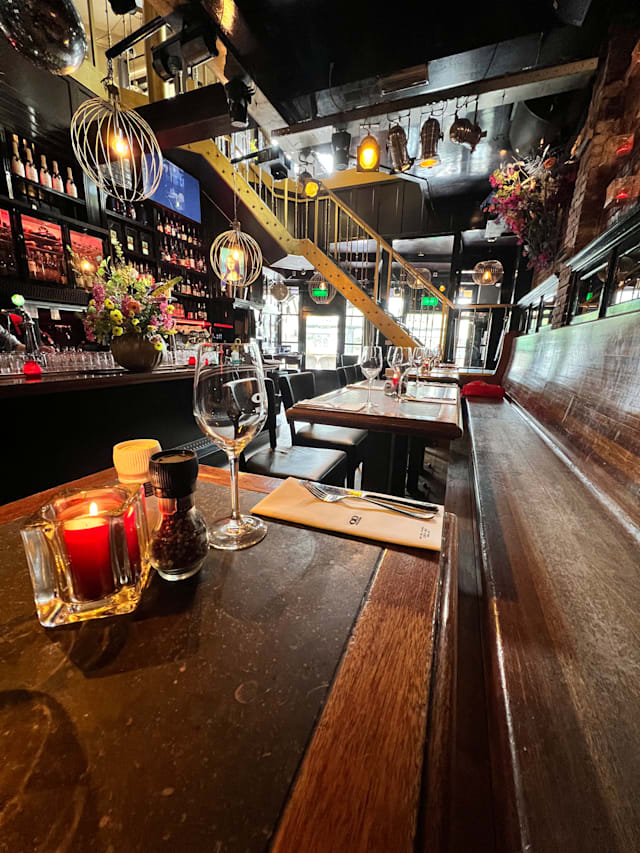 Een stijlvol restaurantinterieur met een mix van moderne en rustieke elementen, verlicht door unieke hanglampen. Een lange houten bar met elegante glazen en een kaars op de tafel. De muren zijn versierd met kleurrijke bloemstukken en planken vol flessen, wat karakter toevoegt aan de sfeer. De zithoek heeft comfortabele banken en tafels, wat een uitnodigende ambiance creëert voor gasten.