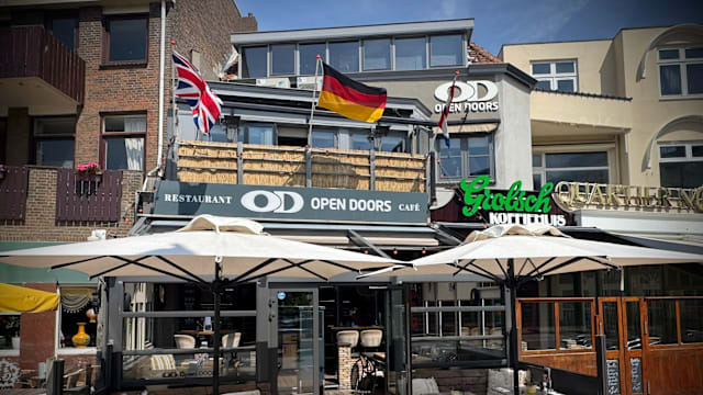 Een goed verlicht restaurant met de naam 'OPEN DOORS' prominent weergegeven. Het gebouw heeft een modern ontwerp met grote ramen en een schone gevel. Vlaggen van het VK en Duitsland wapperen boven de ingang, wat een internationaal tintje toevoegt. Buiten zitplaatsen zijn onder grote parasols gerangschikt, wat een gastvrije ruimte voor gasten creëert.
