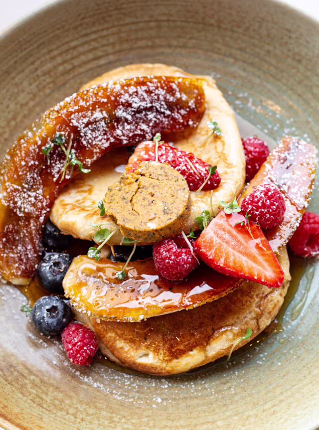 Fluffy pancakes topped with fresh raspberries, strawberries, blueberries, and caramelized banana, drizzled with syrup and finished with a piece of cookie dough and a dusting of powdered sugar.