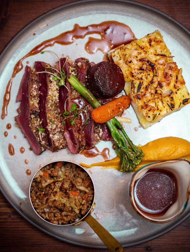 Duck slices with a crispy herb crust, accompanied by chargrilled carrot, asparagus, beetroot, potato gratin, orange puree, and a grain pilaf.