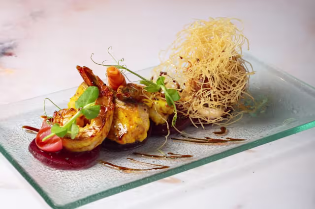 Grilled shrimp served with a rich sauce, garnished with microgreens and vibrant tomatoes, accompanied by crispy fried noodles.