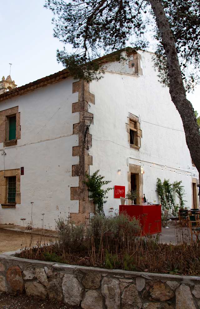 Exterior rústico de un restaurante con edificio de piedra encalada, marcos de ventanas de madera verdes y un letrero rojo que dice 'L'EMP', rodeado de árboles y un jardín que crea un ambiente natural y acogedor.