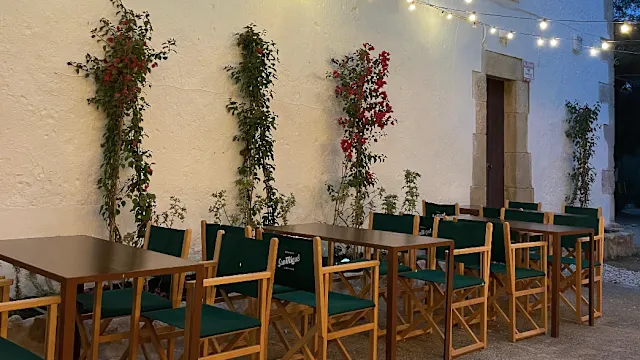 Área de comedor al aire libre con mesas de madera y sillas acolchadas verdes, decorada con plantas trepadoras y flores vibrantes, iluminada con luces suaves.