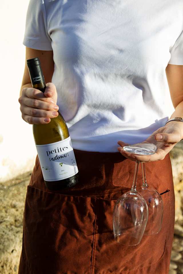 Persona sosteniendo una botella de vino blanco 'petites retours' y un plato pequeño con dos copas de vino, en un ambiente al aire libre.