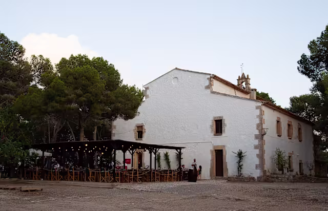 Fachada encantadora de un restaurante rústico con paredes encaladas, techado inclinado y vigas de madera, rodeado de árboles altos y un área de comedor al aire libre bajo una pérgola iluminada.