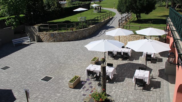 Area esterna del ristorante con tavoli eleganti sotto ombrelloni bianchi, circondata da un paesaggio curato con erba verde e alberi, creando un'atmosfera serena.