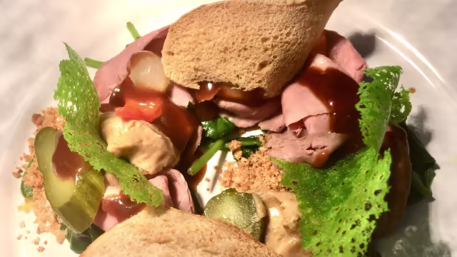 Fette di manzo arrosto tenero con salsa cremosa, cetrioli croccanti, pomodori a cubetti e verdure fresche, guarnite con elementi verdi croccanti e pane sbriciolato.