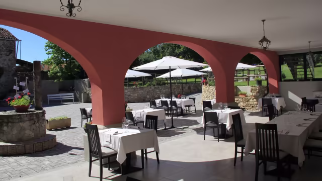 Area esterna di un ristorante con tavoli bianchi e sedie nere, archi eleganti e ombrelloni per riparo, circondata da vegetazione lussureggiante e un muro di pietra.