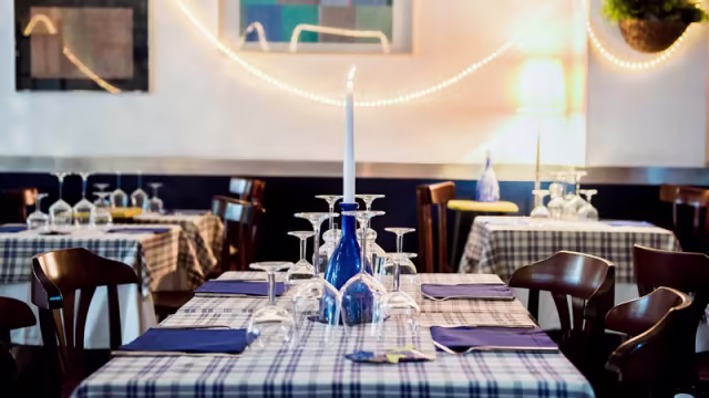 Interno di un ristorante accogliente con tavoli apparecchiati, tovaglie a quadri blu e bianche, bicchieri eleganti e candele bianche, creando un'atmosfera invitante.