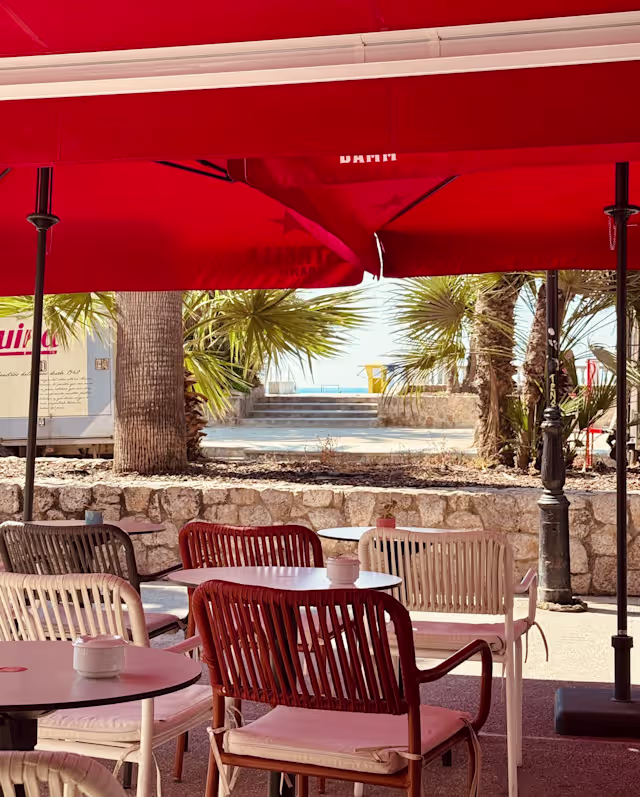 Área de comedor al aire libre con paredes de piedra blanca, muebles de madera y sombrillas rojas, rodeada de palmeras, creando un ambiente tropical y relajado.