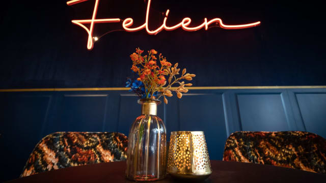 Stijlvol restaurantinterieur met een donkerblauwe muur en een neonreclame "Felien", aangevuld met een vaasje met gedroogde bloemen in warme tinten en een decoratieve gouden kaarsenhouder.