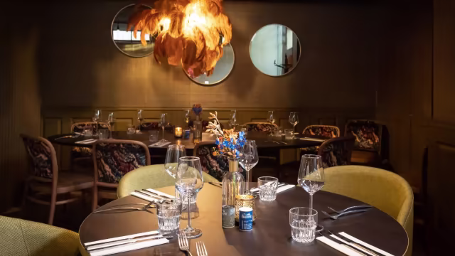 Gezellige restaurantinterieur met een warme ambiance, ronde tafel omringd door elegant gestoffeerde stoelen, versierd met glaswerk en een bloemstuk als centerpiece.