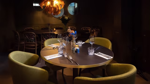 Gezellige restaurantinterieur met een ronde tafel, elegant gedekt met bestek en glazen, omringd door comfortabele groene stoelen en sfeervolle verlichting.