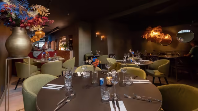 Het restaurantinterieur heeft een moderne en elegante uitstraling met een ronde tafel omringd door luxe groene stoelen en sfeervolle verlichting die een warme ambiance creëert. Decoratieve elementen omvatten een grote, artistieke bloemenarrangement in een gouden vaas en spiegels die de algehele sfeer verbeteren.