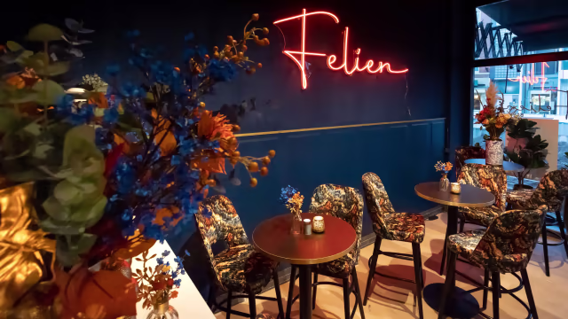 Gezellige restaurantinterieur met een moderne esthetiek, gekenmerkt door een neonbord met de tekst "Felien", omringd door tafels met kleine bloemstukken in blauw en oranje, en gestoffeerde stoelen die bijdragen aan een levendige sfeer.