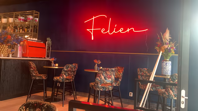 Stijlvol restaurantinterieur met een gezellige sfeer, verlicht door een neonbord "Felien". Hoge stoelen met een kleurrijk bloemenpatroon rond kleine tafels en een moderne bar met decoratieve elementen.