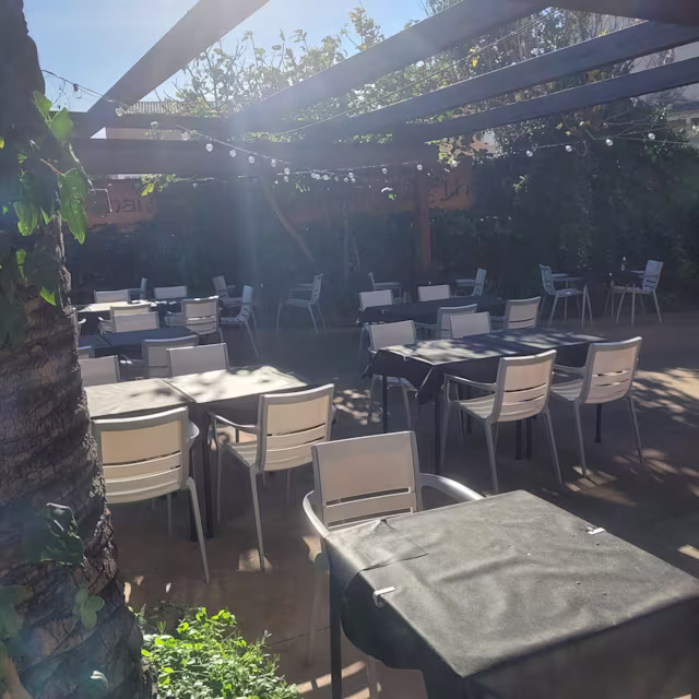 Área de comedor al aire libre con mesas bajo una pérgola de madera, rodeada de vegetación exuberante y con una atmósfera cálida y acogedora.