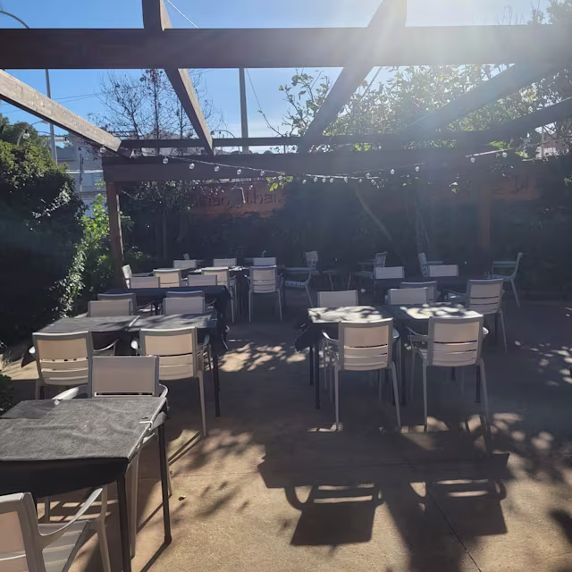 Área de comedor al aire libre con patio espacioso, mesas y sillas blancas, pérgola de madera, vegetación abundante y luces de cadena para una atmósfera acogedora.