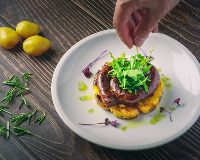 Salsiccia grigliata su un letto cremoso di purè, guarnita con microgreens e un filo di salsa verde.