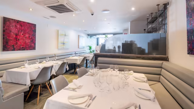 Interior moderno y minimalista con asientos grises, manteles blancos y arte abstracto en tonos rojos y azules, creando un ambiente acogedor y elegante.