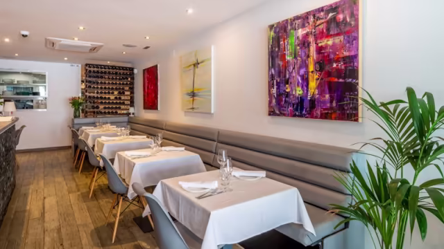 Interior moderno y acogedor con paredes claras, arte abstracto, bancos tapizados grises y mesas de madera minimalistas. Suelo de madera cálida y una pequeña planta añaden un toque de frescura.