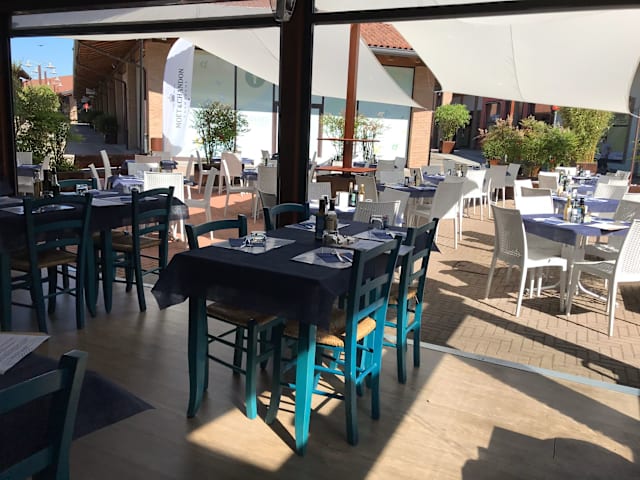 Area esterna del ristorante con tavoli e sedie in un ambiente informale, decorata con tovaglie blu e piante in vaso, creando un'atmosfera accogliente e luminosa.