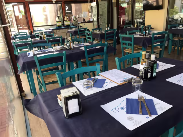 Interno di un ristorante con tavoli apparecchiati, tovaglie blu scuro e sedie turchesi, creando un'atmosfera accogliente e informale.