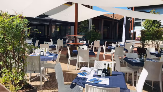 Area esterna di un ristorante con tavoli e sedie bianchi, tovaglie blu scuro e ombrelloni, creando un'atmosfera rilassante e accogliente.