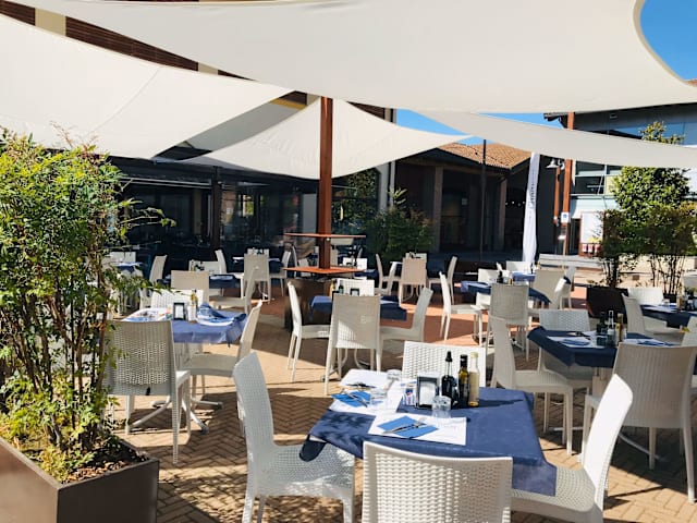 Area esterna del ristorante con tavoli sotto grandi tende bianche, tovaglie blu scuro e piante in vaso, creando un'atmosfera rilassata e accogliente.