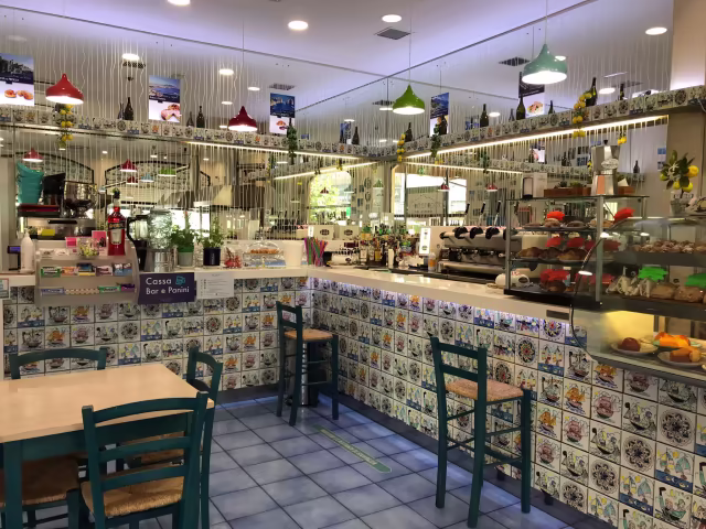 Interno di un ristorante accogliente con un'atmosfera vivace, caratterizzato da un bancone con una selezione di dolci e torte colorate, una macchina da caffè e pareti decorate con piastrelle. L'illuminazione è fornita da lampade a sospensione colorate, mentre l'area dei posti a sedere è composta da semplici tavoli e sedie in legno.