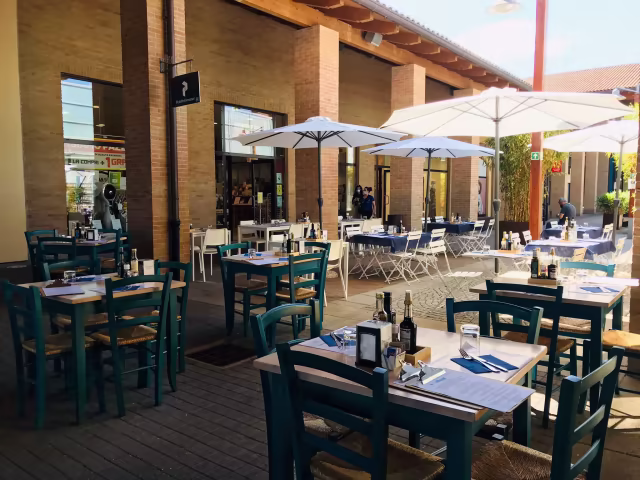 Area esterna del ristorante con tavoli sotto ombrelloni, tovaglioli blu e menù, sedie teal e un'atmosfera vivace.