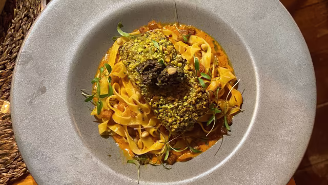 Uma generosa porção de fettuccine com um rico molho cremoso de tomate ou pimento assado, coberta com um pedaço de carne, possivelmente frango ou cordeiro, crocante com pistácios picados e guarnecida com microverdes.