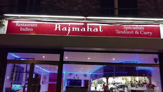 L'extérieur du restaurant indien "Rajmahal" présente un nom en lettres blanches sur fond rouge, mettant en avant ses spécialités tandoori et curry, avec un éclairage bleu doux créant une atmosphère accueillante.