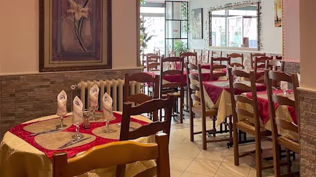 Intérieur du restaurant chaleureux et accueillant, avec des chaises et tables en bois, nappes crème et accents rouges, éclairage naturel et œuvres d'art élégantes.