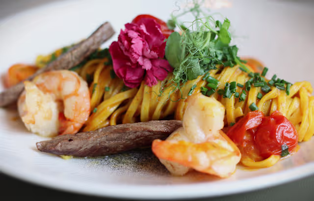 Elegantes Pasta-Gericht mit goldenen Fettuccine, saftigen Garnelen und zartem gegrilltem Rindfleisch, garniert mit frischen Kräutern, einer zarten rosa Blume und Kirschtomaten.