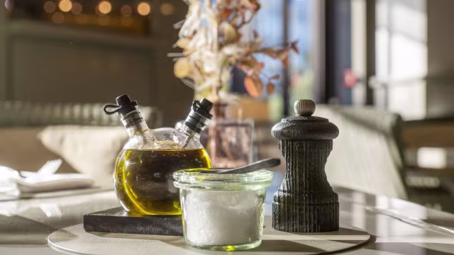 Das Bild zeigt eine stilvolle Tischdekoration mit einem Glasbehälter für grobes Salz, einer dunklen Pfeffermühle sowie zwei kleinen Glasflaschen für Olivenöl und Essig.