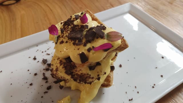 Dessert a strati con fette di pane tostato, ripieno di crema ricca e liscia, decorato con pezzi di cioccolato fondente e petali di fiori edibili.