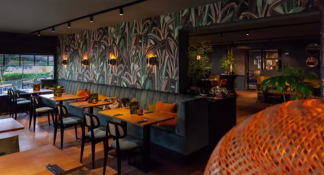 Het restaurant heeft een moderne en uitnodigende inrichting met botanisch behang, comfortabele donkergroene banken en houten tafels, verlicht door zachte wandlampen en voorzien van strategisch geplaatste kamerplanten.