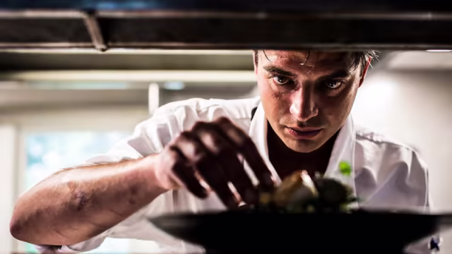 Een chef-kok in een wit uniform bereidt zorgvuldig een gerecht met verschillende ingrediënten, mogelijk vlees of vis, vergezeld van verse kruiden en garneringen, wat vakmanschap en toewijding aan de culinaire kunst uitstraalt.
