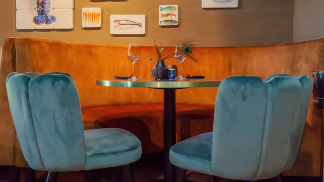 Gezellig restaurantinterieur met een pluche, gebogen bank in warm goudbruin, aangevuld met zachte, teal stoelen. De wanden zijn versierd met ingelijste kunstwerken, waaronder visillustraties, die het aquatische thema versterken.
