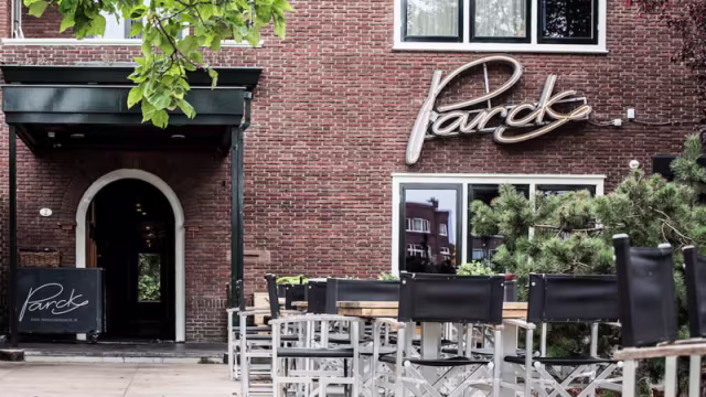 Het exterieur van restaurant "Pearck" met een warme bakstenen gevel, elegante naam in sierlijk schrift, en moderne buitenstoelen, omringd door groen voor een uitnodigende sfeer.