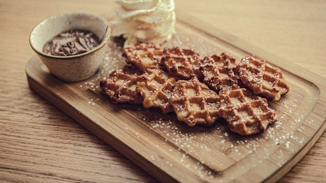 Assiette de gaufres dorées, croustillantes à l'extérieur et moelleuses à l'intérieur, saupoudrées de sucre glace, accompagnées d'un bol de sauce au chocolat et d'une touche de crème fouettée.