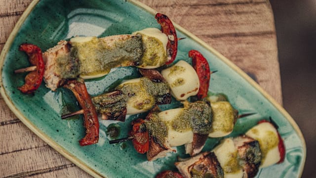 Assiette de brochettes de viande tendre, probablement de poulet, accompagnées de poivrons colorés et d'oignons, le tout arrosé d'une sauce aux herbes vertes et garni de poivrons rouges rôtis.