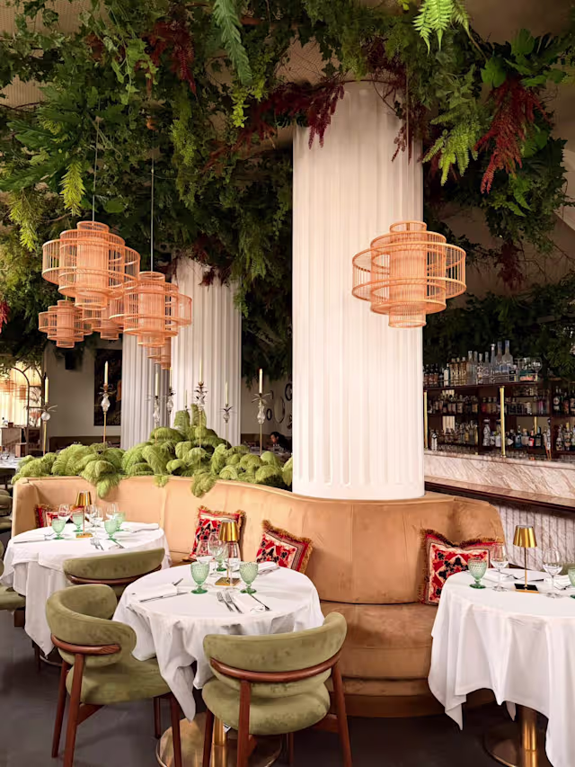 Interni eleganti con vegetazione sospesa, luci a sospensione intrecciate e booth circolari in beige, accompagnati da sedie in velluto verde.
