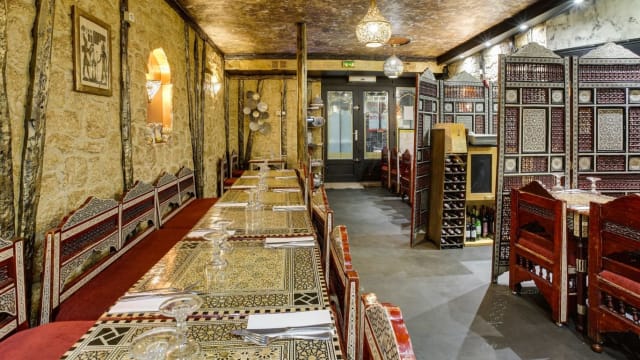 Intérieur du restaurant avec une longue table ornée de motifs traditionnels, des chaises en bois sculptées, des murs texturés ressemblant à de la pierre et un éclairage chaleureux, créant une ambiance conviviale et culturelle.