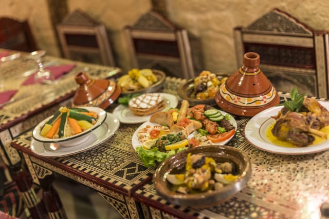 Variété de plats marocains, mettant en avant un tagine traditionnel avec de la viande tendre, probablement de l'agneau, accompagné d'une salade colorée et de brochettes grillées, servies avec du couscous moelleux et des pâtisseries farcies.