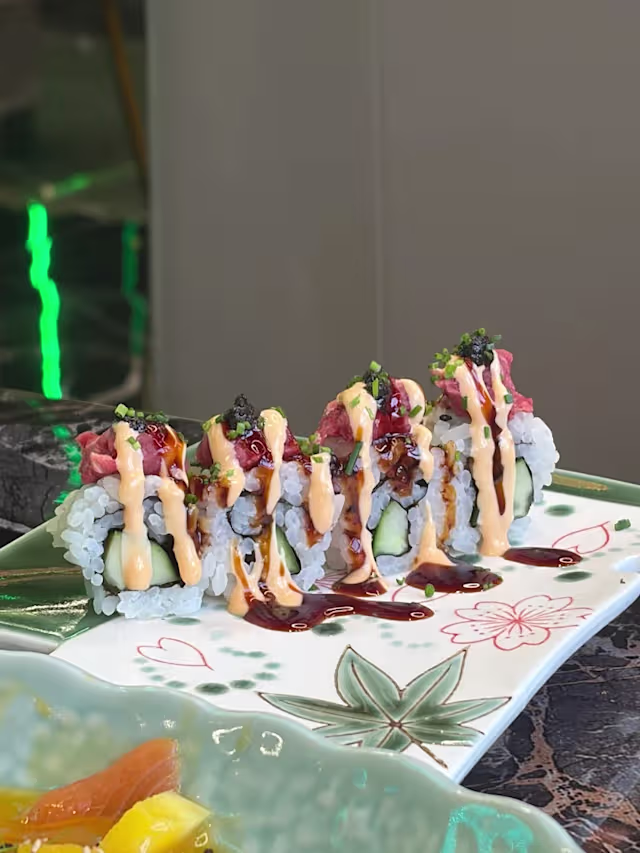 Rouleau de sushi joliment présenté avec des couches de poisson frais, probablement du thon, sur un lit de riz et d'algues, arrosé d'une sauce crémeuse et d'un glaçage sucré, garni d'herbes vertes finement hachées.