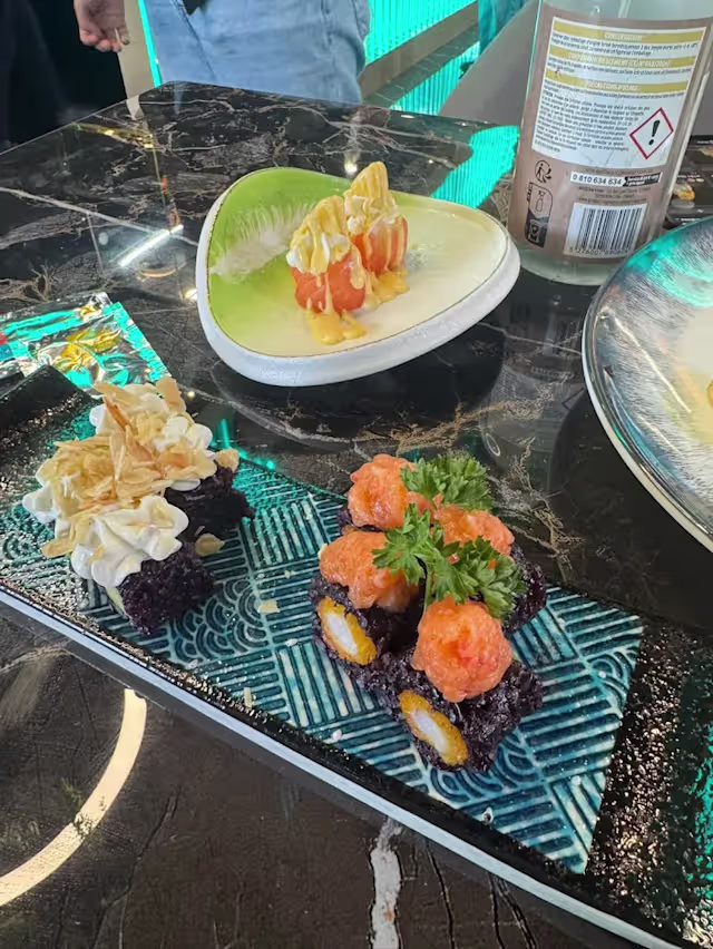 Plateau de sushi joliment présenté avec une variété de rouleaux colorés, dont l'un est garni d'œufs de poisson orange et d'une herbe verte, tandis qu'un autre rouleau est décoré de flocons croustillants.